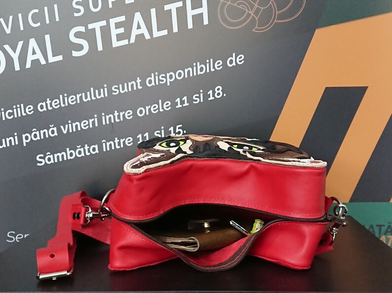 May include: A red leather crossbody bag with a flap featuring an embroidered cat face. The bag is open, revealing a brown leather wallet and other items inside.