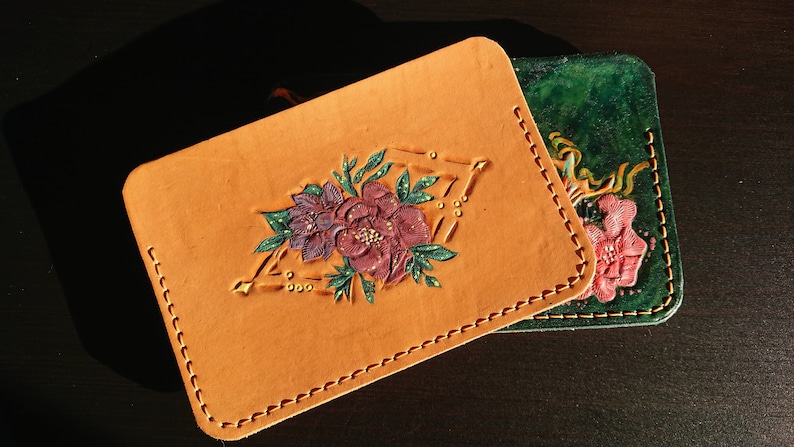 May include: Two leather wallets with floral designs. The top wallet is tan with a pink and purple flower design. The bottom wallet is green with a pink flower design.