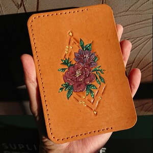 May include: A brown leather card holder with a geometric design and a floral motif in shades of purple, pink, and green. The design features a diamond shape with a floral bouquet inside.