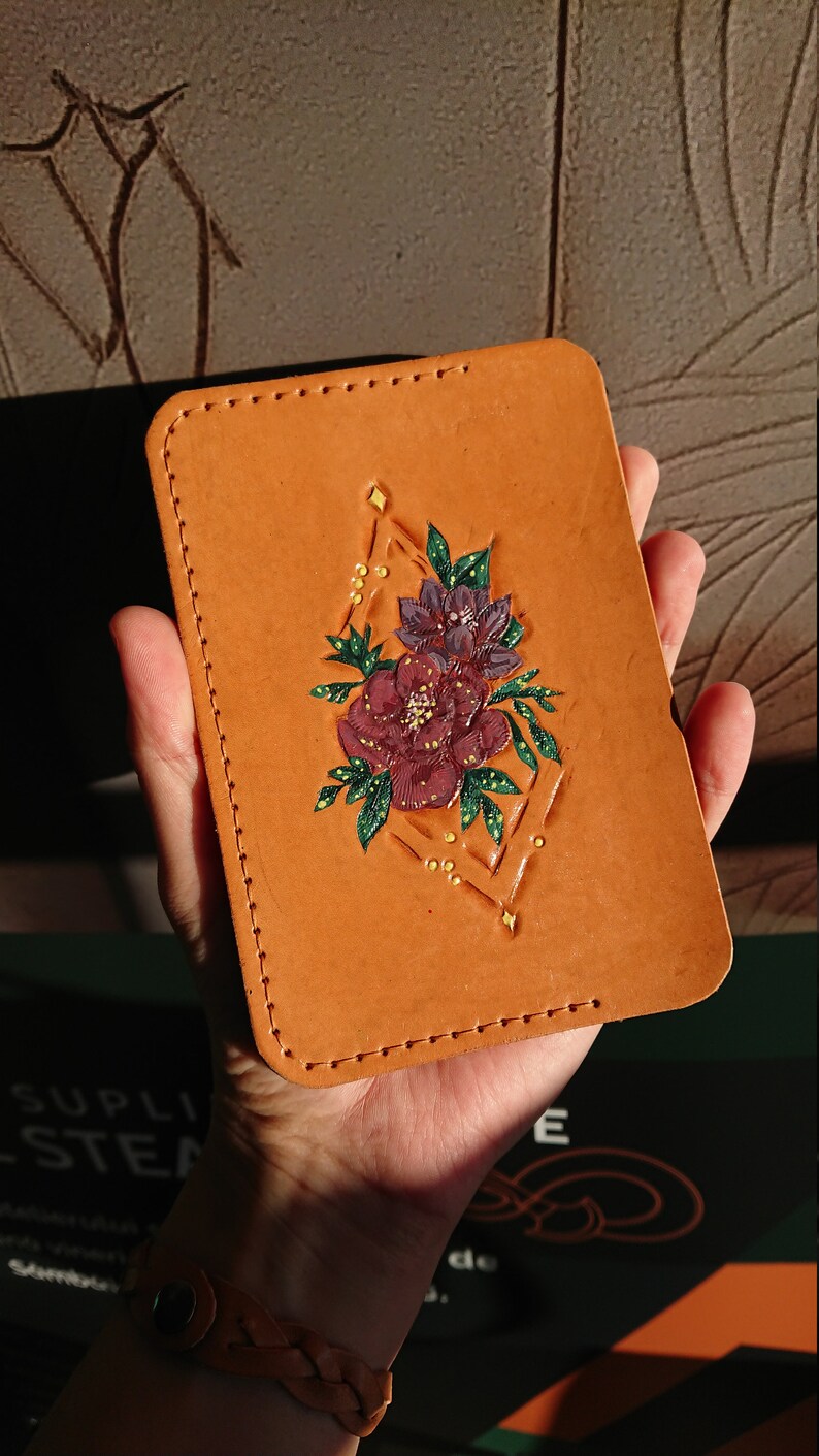May include: A brown leather card holder with a geometric design and a floral motif in shades of purple, pink, and green. The design features a diamond shape with a floral bouquet inside.