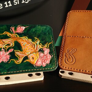 May include: Two leather wallets, one with a green background and a gold fish with pink flowers, the other is brown with a stitched design.