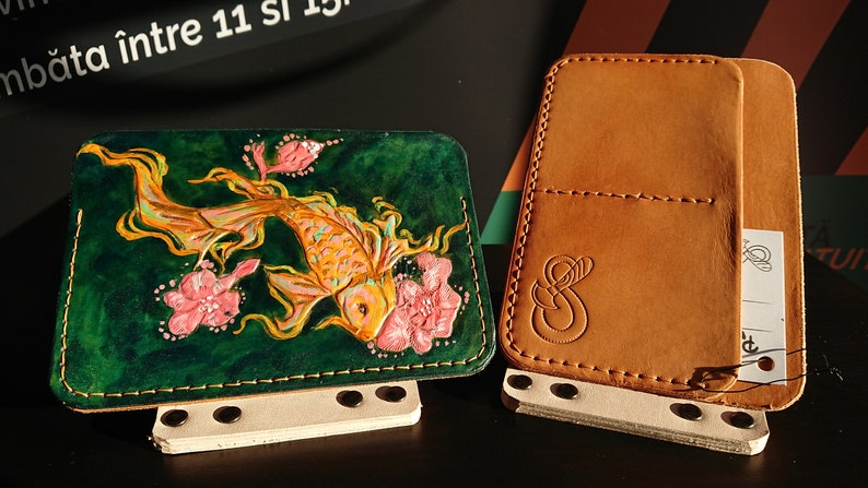 May include: Two leather wallets, one with a green background and a gold fish with pink flowers, the other is brown with a stitched design.