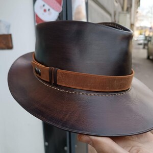 Może przedstawiać: Brązowy sk&oacute;rzany kapelusz fedora z brązowym sk&oacute;rzanym paskiem i szwami. Kapelusz ma szeroki rondel i wysoki korpus.