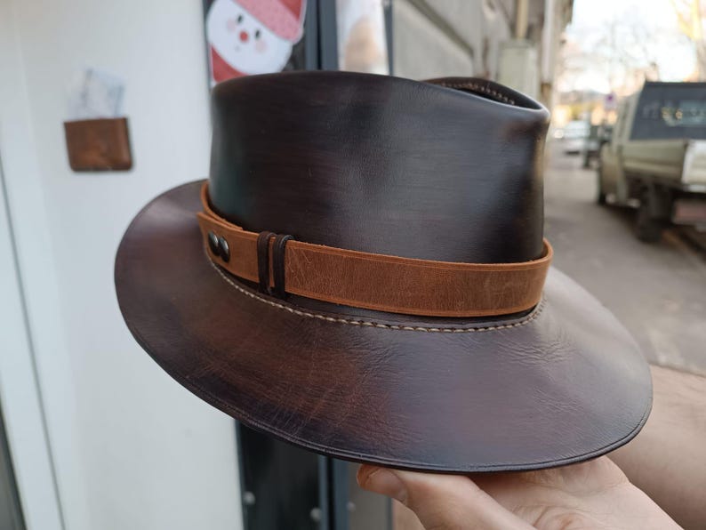Może przedstawiać: Brązowy sk&oacute;rzany kapelusz fedora z brązowym sk&oacute;rzanym paskiem i szwami. Kapelusz ma szeroki rondel i wysoki korpus.