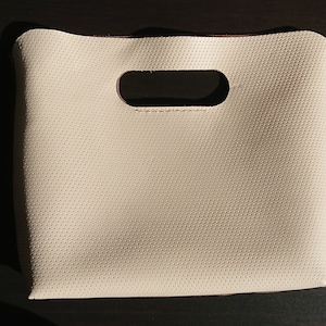 Puede incluir: Una pequeña bolsa de mano de color blanco roto con un asa recortada. La bolsa está hecha de un material texturizado con pequeños agujeros en toda la superficie.