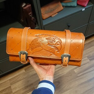 May include: A brown leather motorcycle tool roll with an eagle design embossed on the front. The roll has two buckles and a quilted pattern.