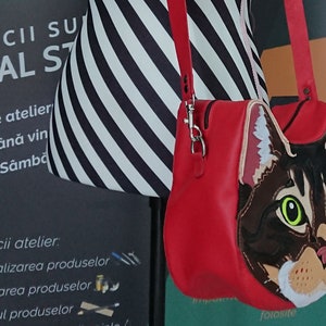 May include: A red leather crossbody bag with a cat face design. The cat has brown fur, green eyes, and a white nose and chin. The bag has a zipper closure and a silver chain strap.