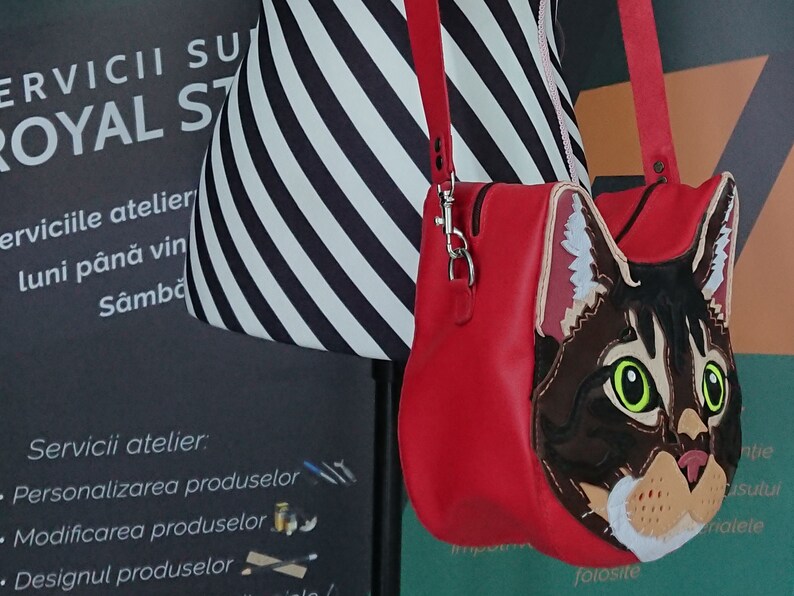 May include: A red leather crossbody bag with a cat face design. The cat has brown fur, green eyes, and a white nose and chin. The bag has a zipper closure and a silver chain strap.