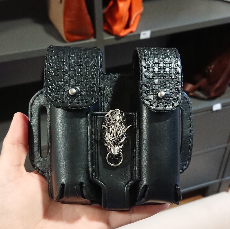 以下が含まれることがあります： 黒革製のデュアルポーチアクセサリー。銀色のオオカミの頭のエンブレムが付いています。ポーチには織り模様と花の模様のアクセントがあります。このアクセサリーは、ベルトに着用するように設計されています。
