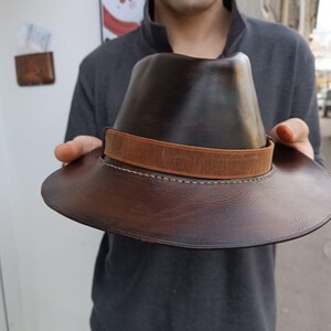 Może przedstawiać: Brązowy sk&oacute;rzany kapelusz fedora z brązowym sk&oacute;rzanym paskiem i szwami. Kapelusz ma szeroki rondel.