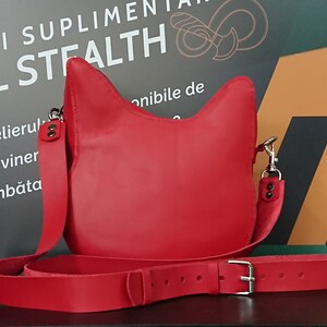 May include: A bright red leather crossbody bag with a cat-shaped flap. The bag has a long adjustable strap and a silver buckle.