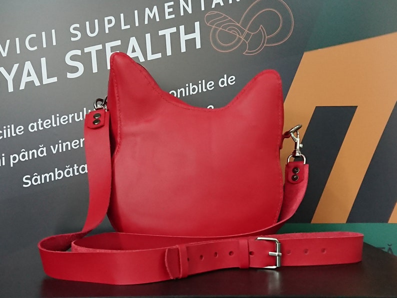 May include: A bright red leather crossbody bag with a cat-shaped flap. The bag has a long adjustable strap and a silver buckle.
