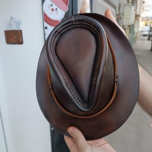 Może przedstawiać: Brązowy sk&oacute;rzany kapelusz fedora z ozdobnym szwem na koronie. Kapelusz ma szeroki rondel i jest wykonany z miękkiej, elastycznej sk&oacute;ry.