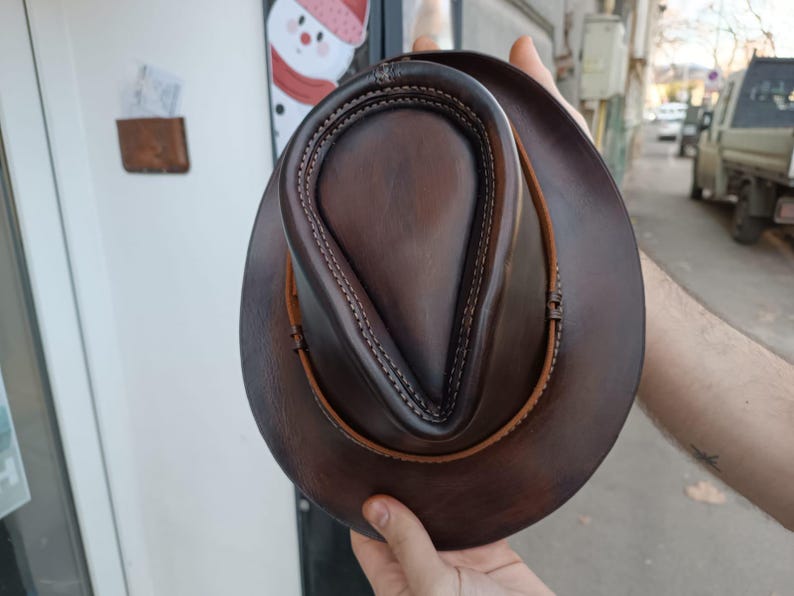 Może przedstawiać: Brązowy sk&oacute;rzany kapelusz fedora z ozdobnym szwem na koronie. Kapelusz ma szeroki rondel i jest wykonany z miękkiej, elastycznej sk&oacute;ry.