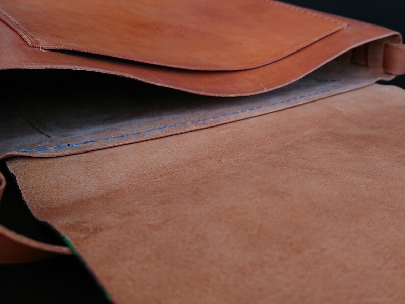 May include: Open view of a brown leather bag with a flap. The interior reveals a lighter brown suede lining and blue stitching. The bag's design suggests a messenger or shoulder bag style.