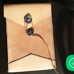 Puede incluir: Una bolsa de estilo sobre de cuero de color beige con dos cierres metálicos decorativos y una larga correa de cuero. La bolsa está cerrada y tiene una solapa cosida.