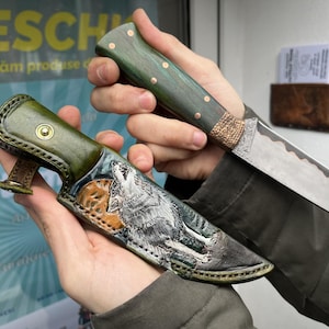May include: A handcrafted hunting knife with a green wooden handle and a decorative blade, held next to a leather sheath. The sheath features a detailed wolf illustration in shades of grey and brown.