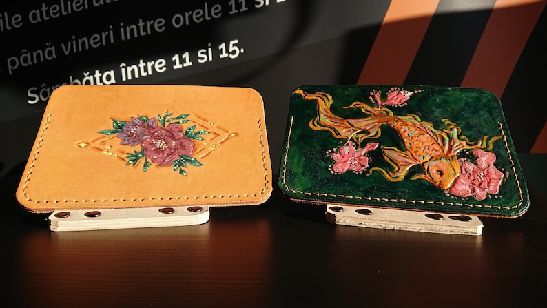 May include: Two leather wallets with hand-painted designs. One wallet is tan with a floral design in shades of pink, purple, and green. The other wallet is green with a gold fish and pink flowers.