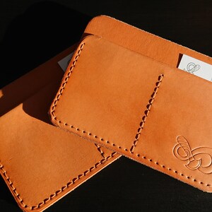 May include: Two brown leather wallets with stitched edges. The top wallet has a small pocket and a larger pocket with a decorative design. The bottom wallet is a simple design with a stitched edge.