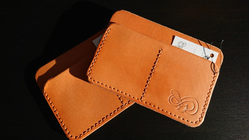 May include: Two brown leather wallets with stitched edges. The top wallet has a small pocket and a larger pocket with a decorative design. The bottom wallet is a simple design with a stitched edge.