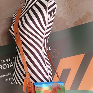 May include: A brown leather shoulder bag with a painted image of a brown horse in a field. The bag has a long adjustable strap and is displayed on a mannequin with black and white stripes.
