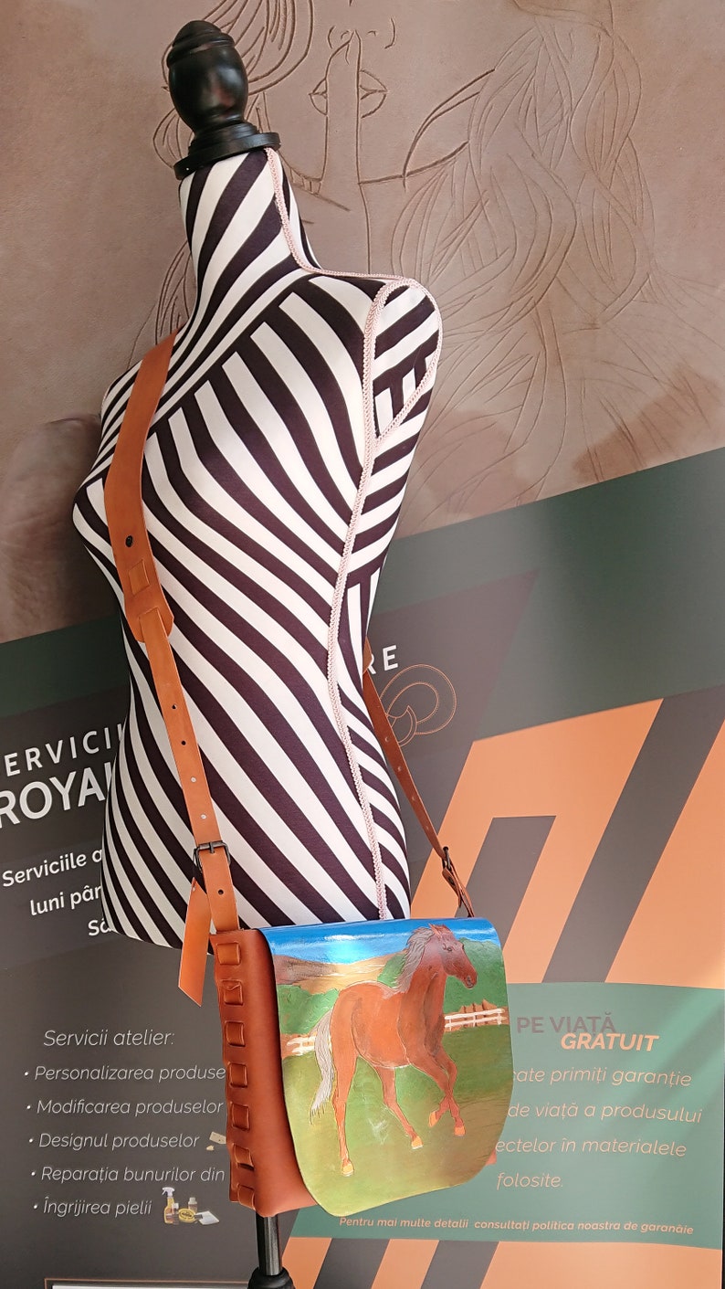 May include: A brown leather shoulder bag with a painted image of a brown horse in a field. The bag has a long adjustable strap and is displayed on a mannequin with black and white stripes.