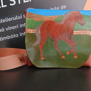 May include: A brown leather crossbody bag with a painted horse scene. The bag features a running horse against a green field, blue sky, and a white fence. Includes a matching leather strap.