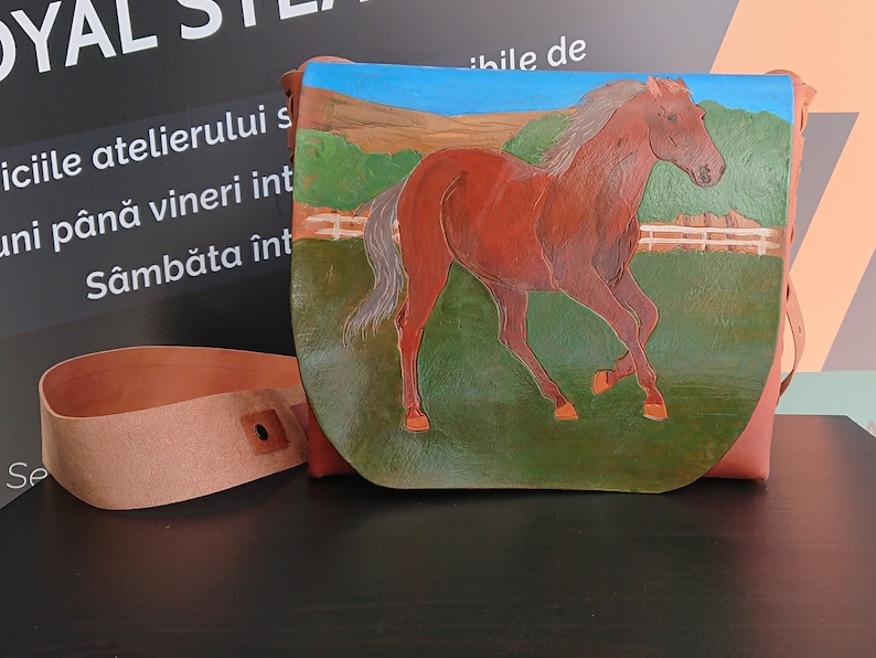 May include: A brown leather crossbody bag with a painted horse scene. The bag features a running horse against a green field, blue sky, and a white fence. Includes a matching leather strap.
