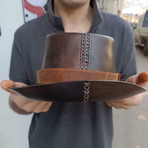 Może przedstawiać: Brązowy sk&oacute;rzany kapelusz fedora z brązowym sk&oacute;rzanym paskiem i detalami szw&oacute;w. Kapelusz jest trzymany w dłoniach osoby.
