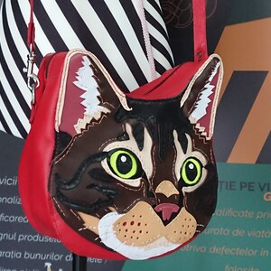 May include: A red leather handbag with a cat face design. The cat's face is made of brown, black, white, and orange leather. The cat has green eyes and a pink nose.