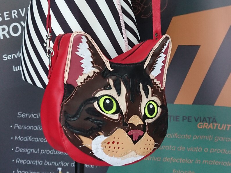 May include: A red leather handbag with a cat face design. The cat's face is made of brown, black, white, and orange leather. The cat has green eyes and a pink nose.