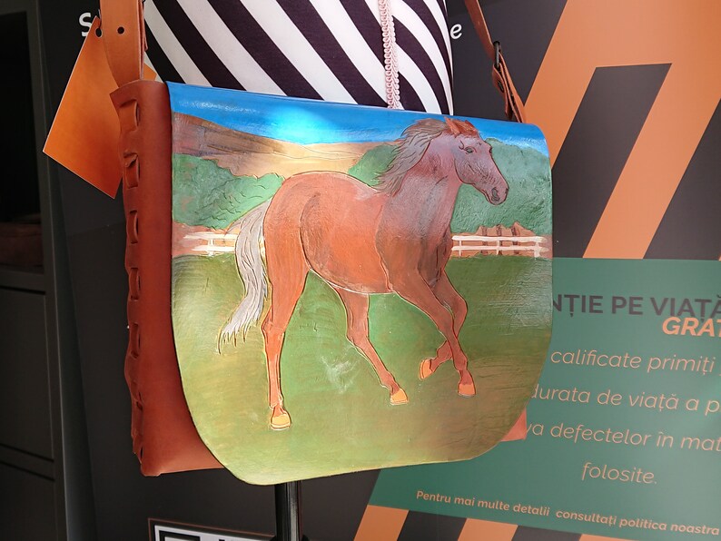 May include: A brown leather shoulder bag with a hand-painted image of a running horse. The horse is brown with a white mane and tail, set against a green field and blue sky. The bag has a brown leather strap.