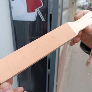 Puede incluir: Una correa de cuero marrón claro con un mango de madera blanco y una correa de cuero beige. La correa se utiliza para afilar navajas de afeitar.