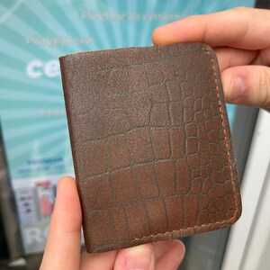 Può includere: Un portafoglio rettangolare in pelle marrone caratterizzato da una texture a rilievo che imita la pelle di coccodrillo sulla superficie esterna. Il portafoglio presenta angoli superiori arrotondati e cuciture visibili lungo i bordi.