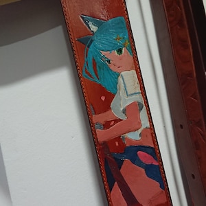 Può includere: Una tracolla per chitarra in pelle marrone con un'immagine dipinta di un personaggio anime con i capelli blu che indossa una camicia bianca e pantaloncini blu.