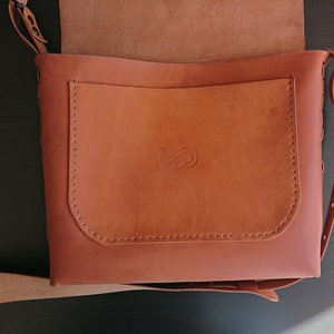 May include: A brown leather shoulder bag with a flap closure and adjustable strap. The bag features a front pocket with a stitched design and a decorative embossed emblem. The bag's dimensions are approximately 12 inches wide and 10 inches tall.