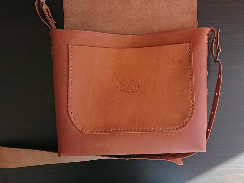 May include: A brown leather shoulder bag with a flap closure and adjustable strap. The bag features a front pocket with a stitched design and a decorative embossed emblem. The bag's dimensions are approximately 12 inches wide and 10 inches tall.