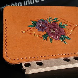 May include: A tan leather wallet with a floral design. The design features a purple and pink flower with green leaves and gold accents.