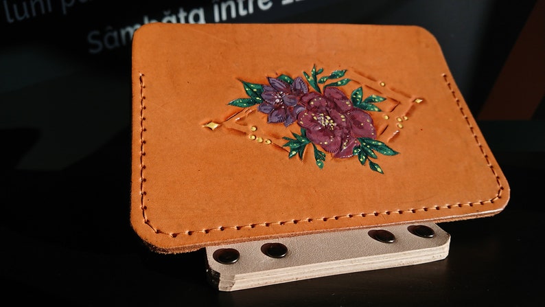 May include: A tan leather wallet with a floral design. The design features a purple and pink flower with green leaves and gold accents.