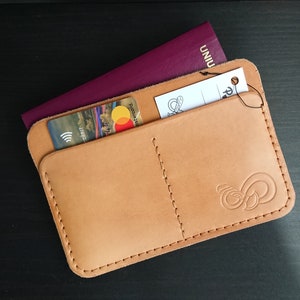 May include: A tan leather passport holder with two pockets, one for a passport and one for cards. A credit card and a passport are visible in the pockets. The holder has a stitched design and a small logo on the bottom right corner.