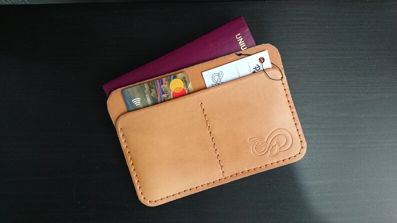 May include: A tan leather passport holder with two pockets, one for a passport and one for cards. A credit card and a passport are visible in the pockets. The holder has a stitched design and a small logo on the bottom right corner.