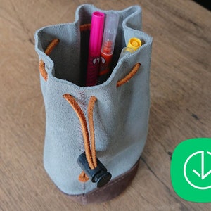 May include: A gray suede drawstring pouch with brown leather trim and a black plastic toggle. The pouch is filled with colorful pens and markers.
