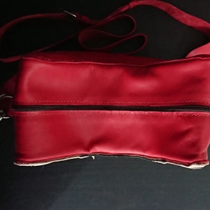 May include: A red leather satchel bag with a zipper closure and a long adjustable strap. The bag has a simple design and is made of high-quality leather.