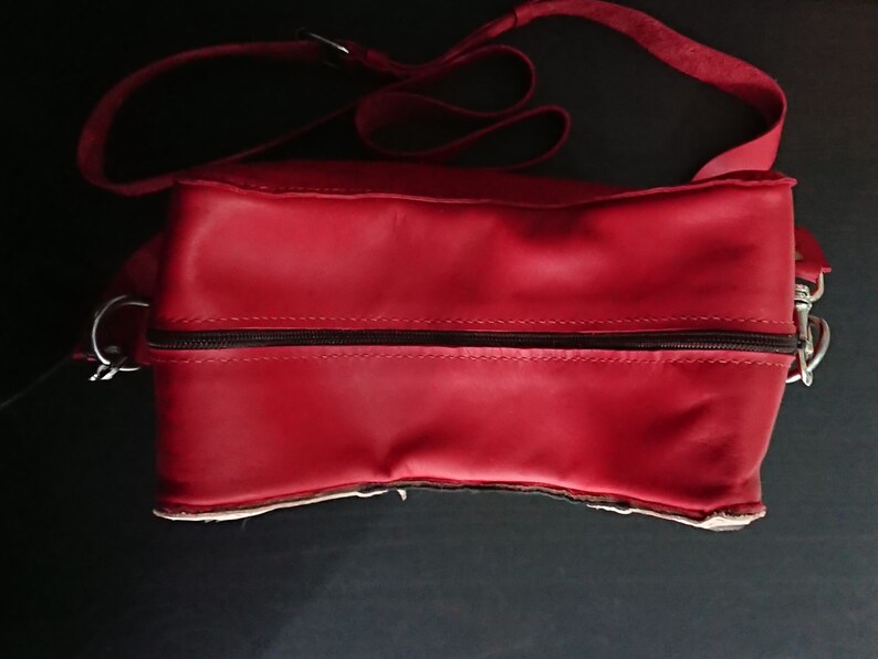 May include: A red leather satchel bag with a zipper closure and a long adjustable strap. The bag has a simple design and is made of high-quality leather.