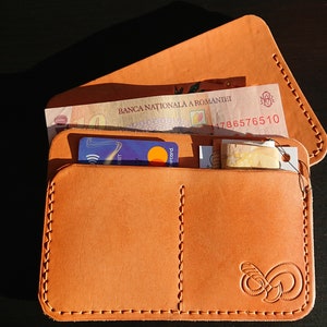 May include: A brown leather wallet with a stitched edge and a small logo on the front. The wallet is open and contains a credit card, cash, and a small piece of paper.