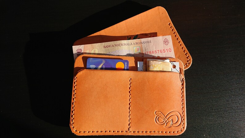 May include: A brown leather wallet with a stitched edge and a small logo on the front. The wallet is open and contains a credit card, cash, and a small piece of paper.