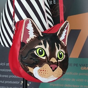 May include: A red leather handbag with a cat face design. The cat's eyes are green, and the fur is brown and black. The bag has a black and white striped strap.