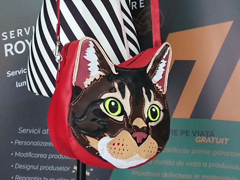 May include: A red leather handbag with a cat face design. The cat's eyes are green, and the fur is brown and black. The bag has a black and white striped strap.