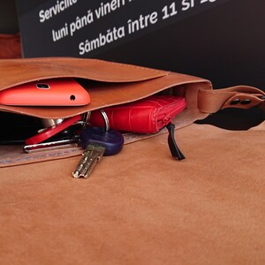May include: Open brown leather bag with a flap, revealing a red phone, keys, and a red wallet. The bag has a strap and is filled with personal items. The bag is made of a soft, textured leather.