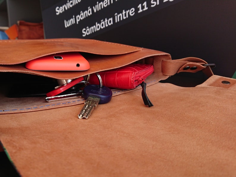 May include: Open brown leather bag with a flap, revealing a red phone, keys, and a red wallet. The bag has a strap and is filled with personal items. The bag is made of a soft, textured leather.